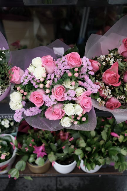 A bouquet of fresh pink roses, white carnations, and light pink statice flowers arranged with green foliage, wrapped in translucent purple paper, and placed on a dark surface at a flower shop, showcas