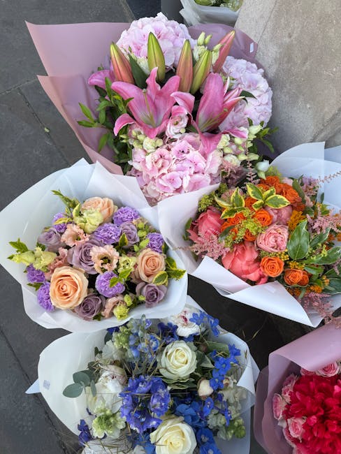 A collection of five vibrant floral bouquets, each wrapped in white or pink paper, arranged on a grey surface. The first bouquet features pink lilies and pale pink hydrangeas, with blooms partially op