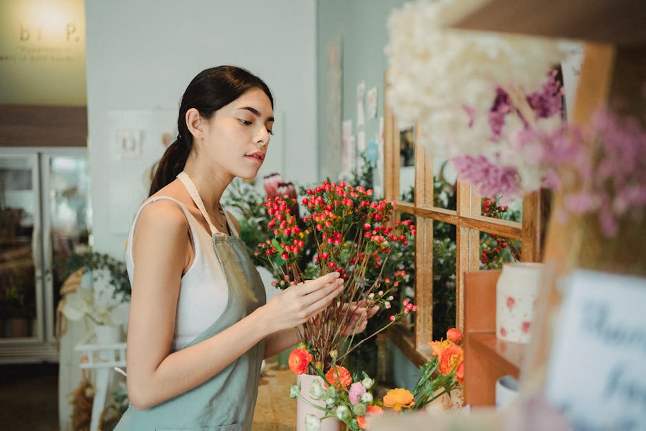 A young woman with dark hair tied back, wearing a white sleeveless top and light grey apron, carefully examining a small bouquet of pink and white buds with green stems at a flower shop. She is standi
