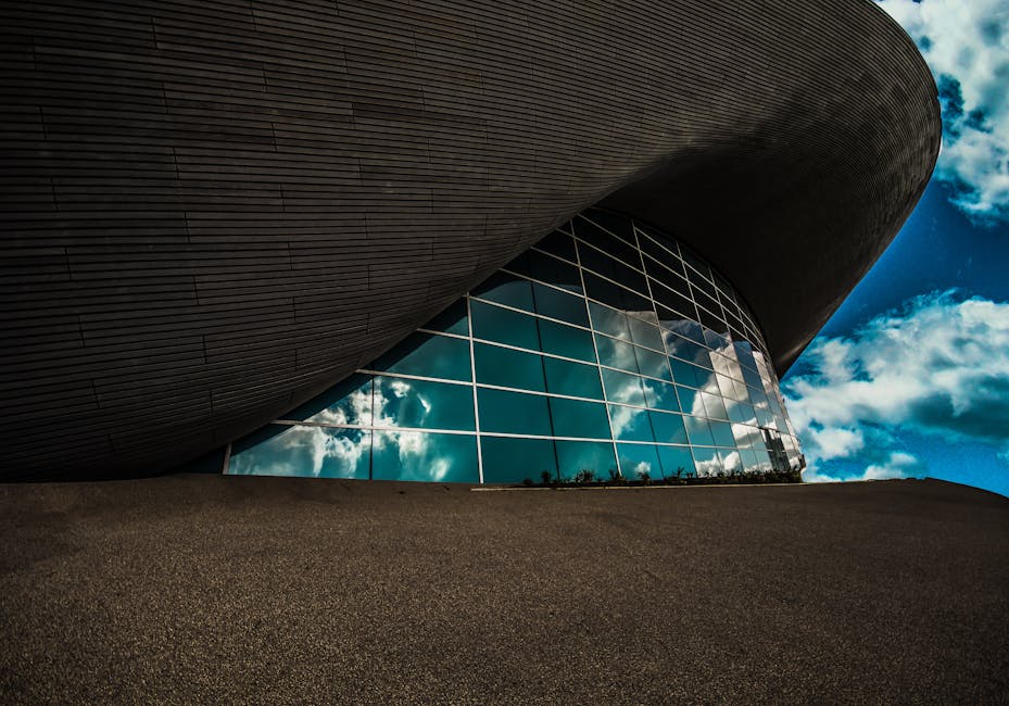 A modern building with a curved, dark exterior and large, reflective glass windows that mirror the blue sky and white clouds above. The structure is set on a textured, sloped surface, with minimal lan